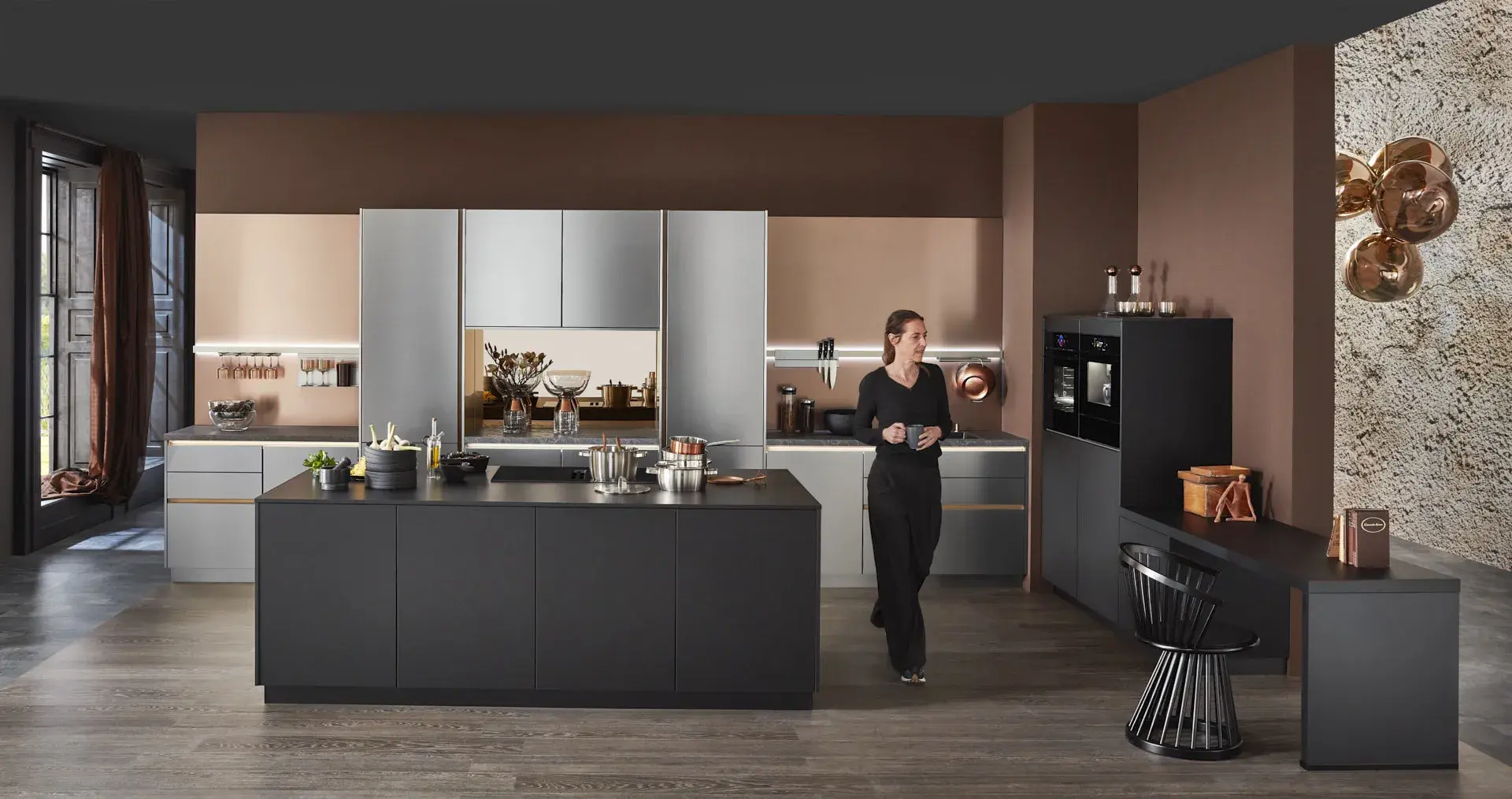 Hauptschuss, Mix aus Keramik und Bronzenachbildung, Kücheninsel mit Bosch Induktionskochfeld und Muldenlüfter, schwarzer Schrank, Edelstahlfronten, Bronze Nischenrückwand, 2 Bosch Einbaubacköfen, Messing Spüle, Model ist neben der Kücheninsel